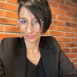 The way to sale - Portret kobiety w czarnej marynarce i okularach, uśmiechniętej, z krótkimi ciemnymi włosami, na tle ceglanej ściany.