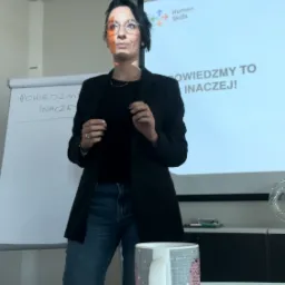 Kobieta w okularach prezentuje slajd z napisem 'Powiedzmy to inaczej!' i logo Human Skills. Na stole kubek. Prezentacja w jasnym pomieszczeniu.