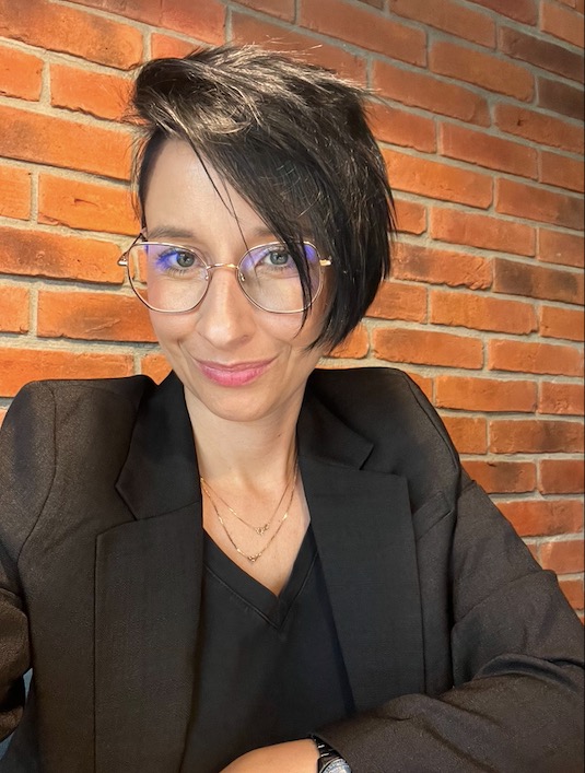 Portret kobiety w czarnej marynarce i okularach, uśmiechniętej, z krótkimi ciemnymi włosami, na tle ceglanej ściany.
