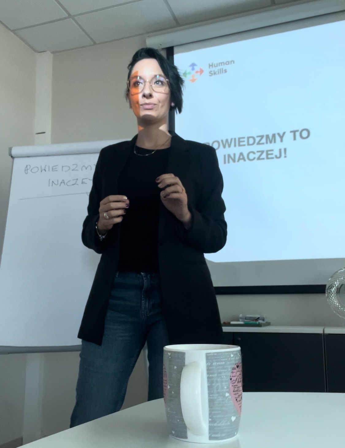 Kobieta w okularach prezentuje slajd z napisem 'Powiedzmy to inaczej!' i logo Human Skills. Na stole kubek. Prezentacja w jasnym pomieszczeniu.
