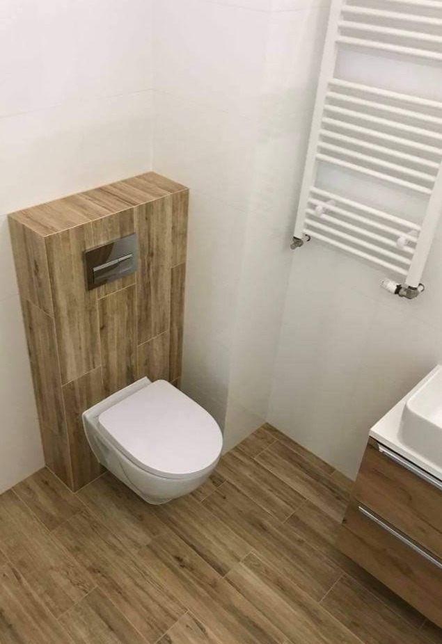 Nowoczesna łazienka z podwieszaną toaletą obudowaną płytkami imitującymi drewno, grzejnikiem drabinkowym i umywalką z szafką w odcieniach drewna.
