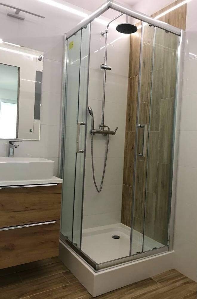 Nowoczesna kabina prysznicowa z deszczownicą i panelami imitującymi drewno, umywalka nablatowa z szafką w odcieniach brązu, lustro z oświetleniem LED w łazience wykończonej białymi płytkami.