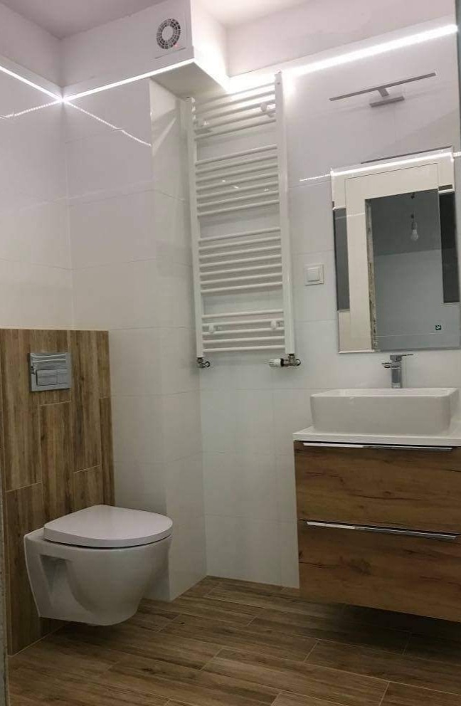 Nowoczesna łazienka z podwieszaną toaletą, drewnopodobnymi płytkami na ścianie i podłodze, białą umywalką nablatową z szafką w kolorze drewna oraz grzejnikiem drabinkowym.