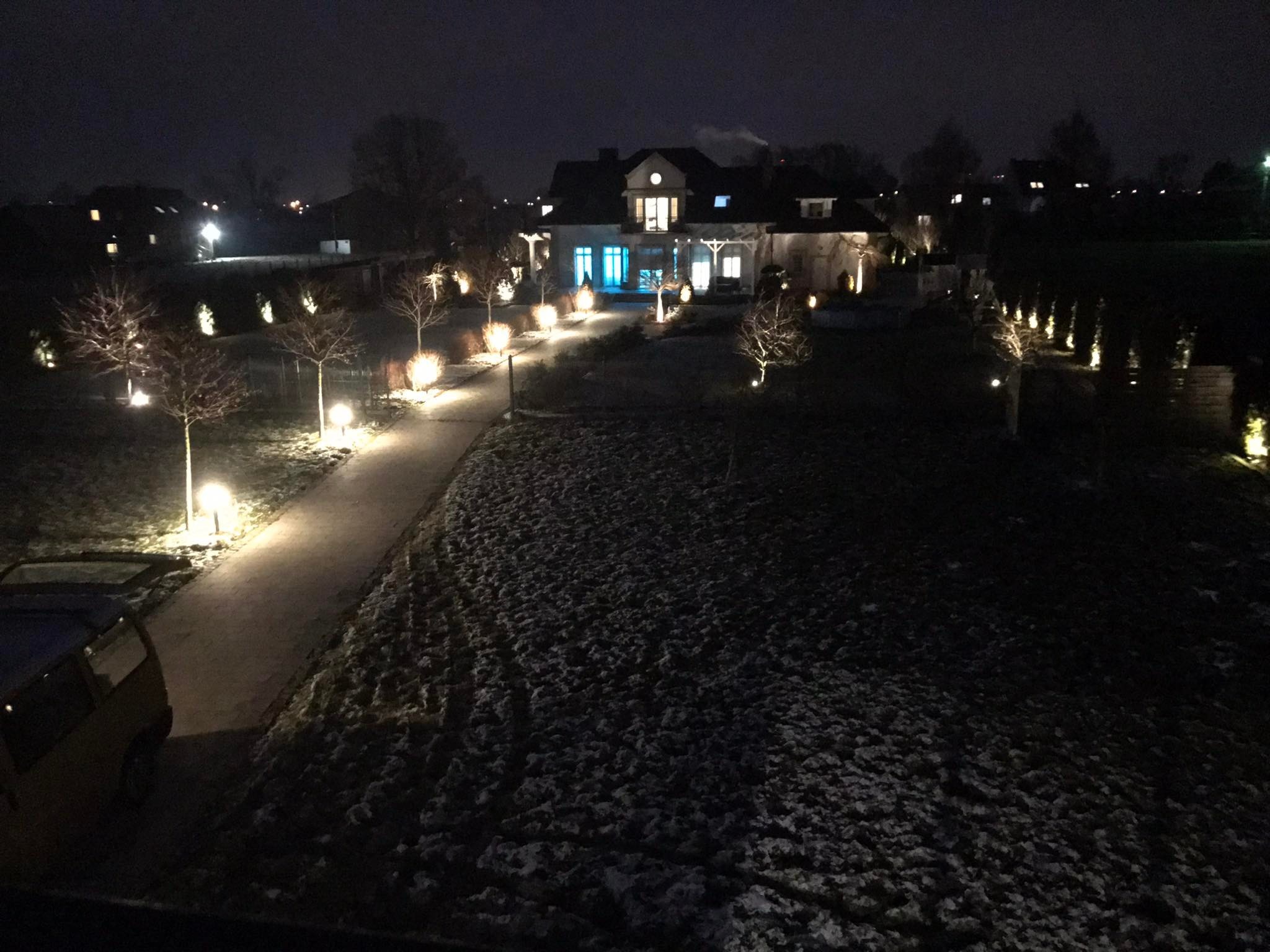 Nocne oświetlenie ogrodowe posesji z podświetlonymi drzewkami wzdłuż ścieżki i elewacją budynku z niebieskim akcentem świetlnym