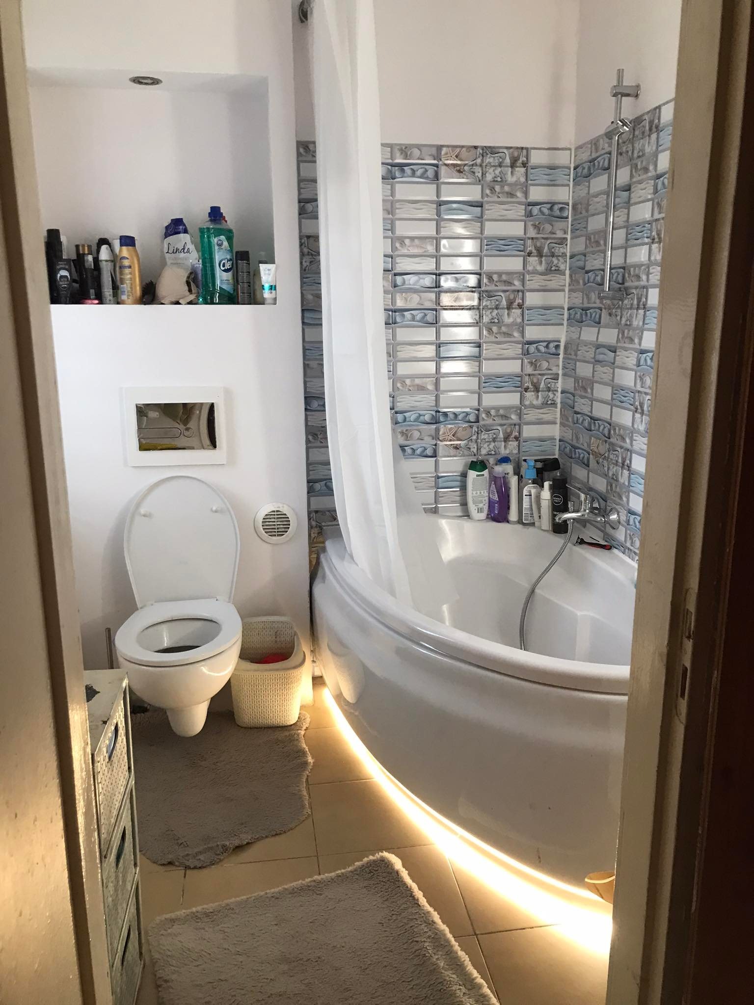 Wnętrze łazienki z podświetloną wanną LED, białą ceramiką i niebiesko-białymi płytkami, widok zza drzwi.