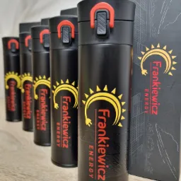 Rząd czarnych kubków termicznych z czerwonymi akcentami i kolorowym nadrukiem firmowym 'Frankiewicz Energy' na jasnym drewnianym blacie, obok czarne pudełko z nadrukiem budynku.