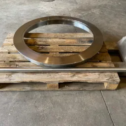 Polerowany, metalowy pierścień i pręt na drewnianej palecie w szarym pomieszczeniu. Widoczne ślady obróbki.