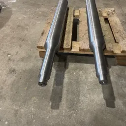 Dwa metalowe, cylindryczne elementy tokarskie na drewnianej palecie w warsztacie. Widoczne ślady obróbki, gładka powierzchnia. Szary beton w tle.