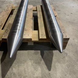 DaMet Damian Napora - Dwa metalowe, stożkowe elementy na drewnianej palecie, o gładkiej, srebrzystej powierzchni. Widoczne ślady obróbki, surowe tło betonowej podłogi.