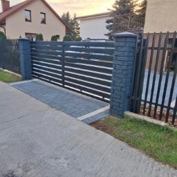 DaMet Damian Napora - Nowoczesna, pozioma brama wjazdowa z czarnymi przęsłami i murowanymi słupkami, uzupełniona o ogrodzenie panelowe z pionowych prętów. Kostka brukowa na podjeździe.