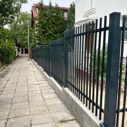 DaMet Damian Napora - Nowoczesne, pionowe ogrodzenie metalowe w kolorze grafitowym, osadzone na betonowej podmurówce, przylegające do chodnika z kostki. W tle dom z czerwoną dachówką.