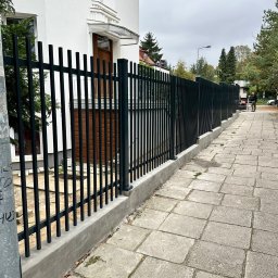 DaMet Damian Napora - Nowoczesne, czarne ogrodzenie metalowe z pionowymi prętami, osadzone na betonowej podmurówce, przylegające do białego budynku z drewnianymi drzwiami wejściowymi.