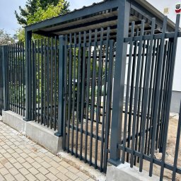 DaMet Damian Napora - Nowoczesna, grafitowa brama wjazdowa z pionowymi szczeblami i zadaszeniem, osadzona na betonowej podmurówce, widok z zewnątrz na tle kostki brukowej i zieleni.