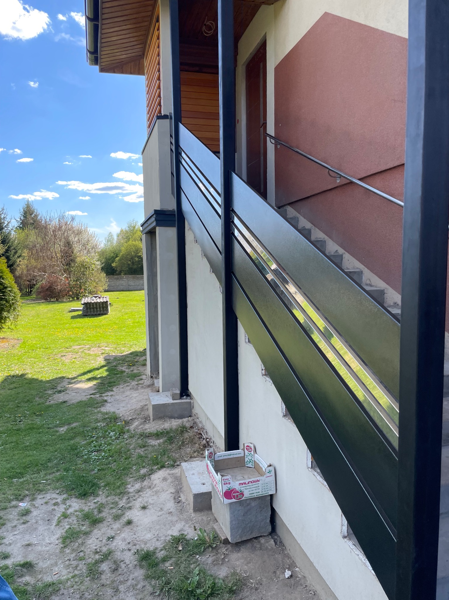 Nowoczesna, czarna balustrada schodowa z poziomymi szczeblami, zamontowana przy zewnętrznych schodach domu z brązową elewacją. W tle widoczny trawnik i drzewa.