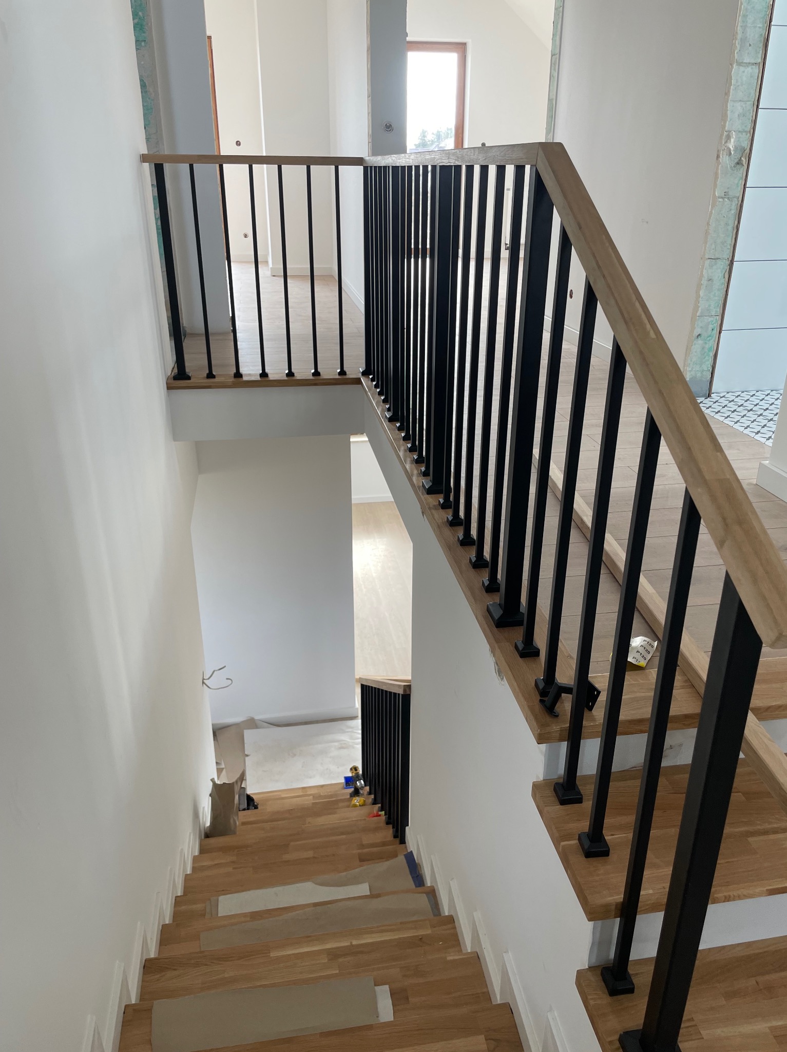 Nowoczesna balustrada schodowa z czarnymi, pionowymi prętami i drewnianym poręczą, widok z góry na drewniane schody w jasnym, minimalistycznym wnętrzu.