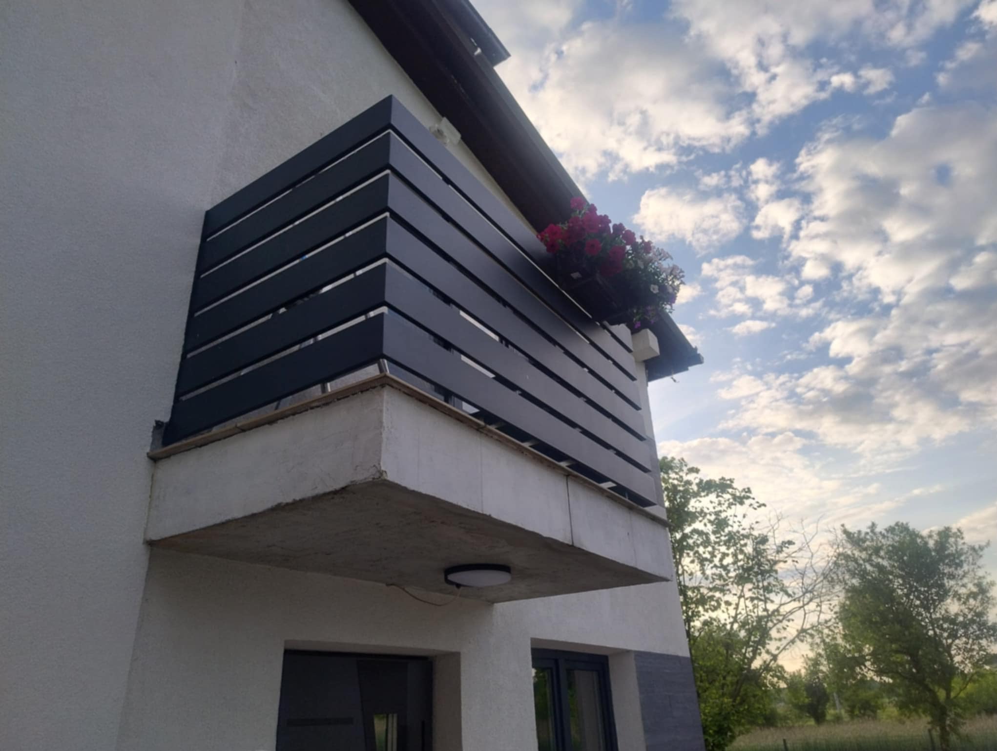 Nowoczesna balustrada balkonowa z poziomych, czarnych profili, uzupełniona kwiatami w skrzynce. Widok z dołu na tle błękitnego nieba.