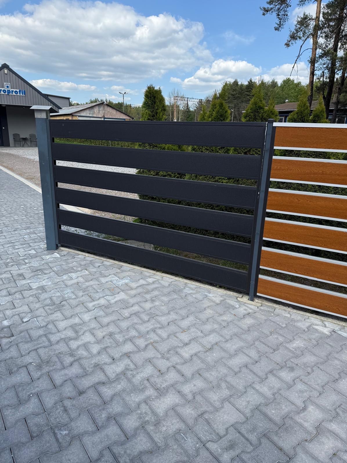 Nowoczesne ogrodzenie panelowe z poziomymi, czarnymi przęsłami i drewnopodobnymi elementami, widoczne na tle kostki brukowej i zieleni. Minimalistyczny design.