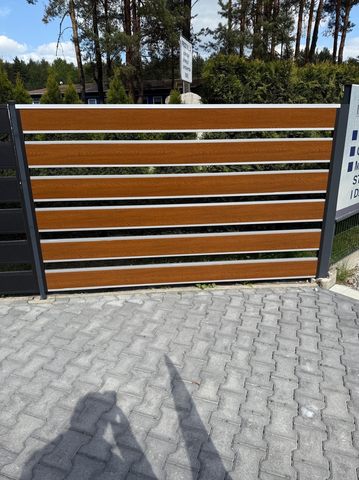 Nowoczesne ogrodzenie poziome z paneli imitujących drewno, z metalowymi słupkami, na tle zieleni. Widoczna kostka brukowa i cień osoby robiącej zdjęcie.