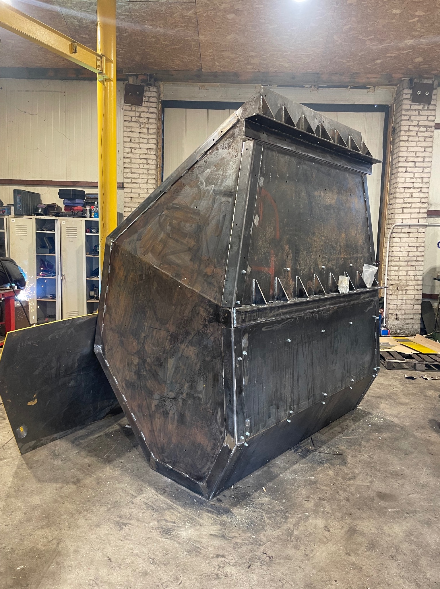 Spawana, surowa konstrukcja metalowa o nietypowym kształcie, z widocznymi spawami i śrubami, stojąca w warsztacie. Element z otwartą klapą z boku. Widoczny sprzęt spawalniczy.