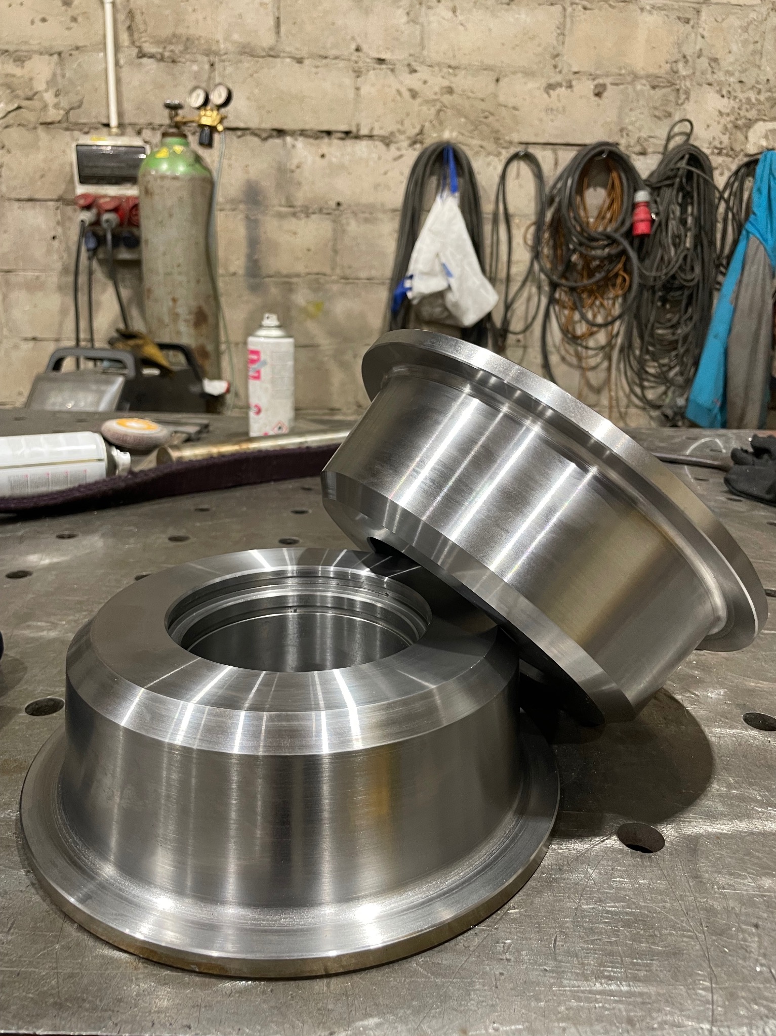 Dwa precyzyjnie obrobione, metalowe pierścienie na warsztatowym stole z otworami, w tle ściana z cegieł i narzędzia ślusarskie. Metaliczny połysk.