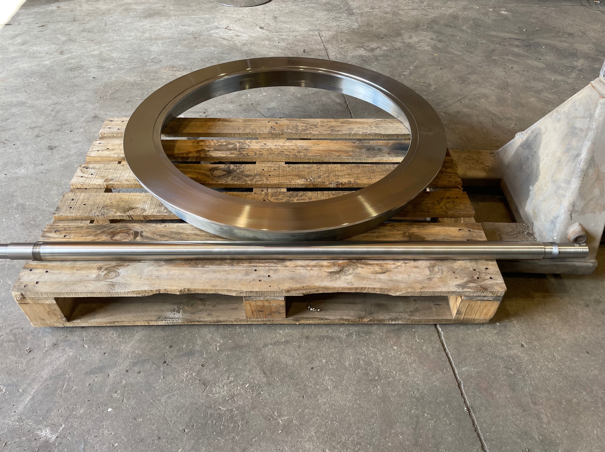 Polerowany, metalowy pierścień i pręt na drewnianej palecie w szarym pomieszczeniu. Widoczne ślady obróbki.