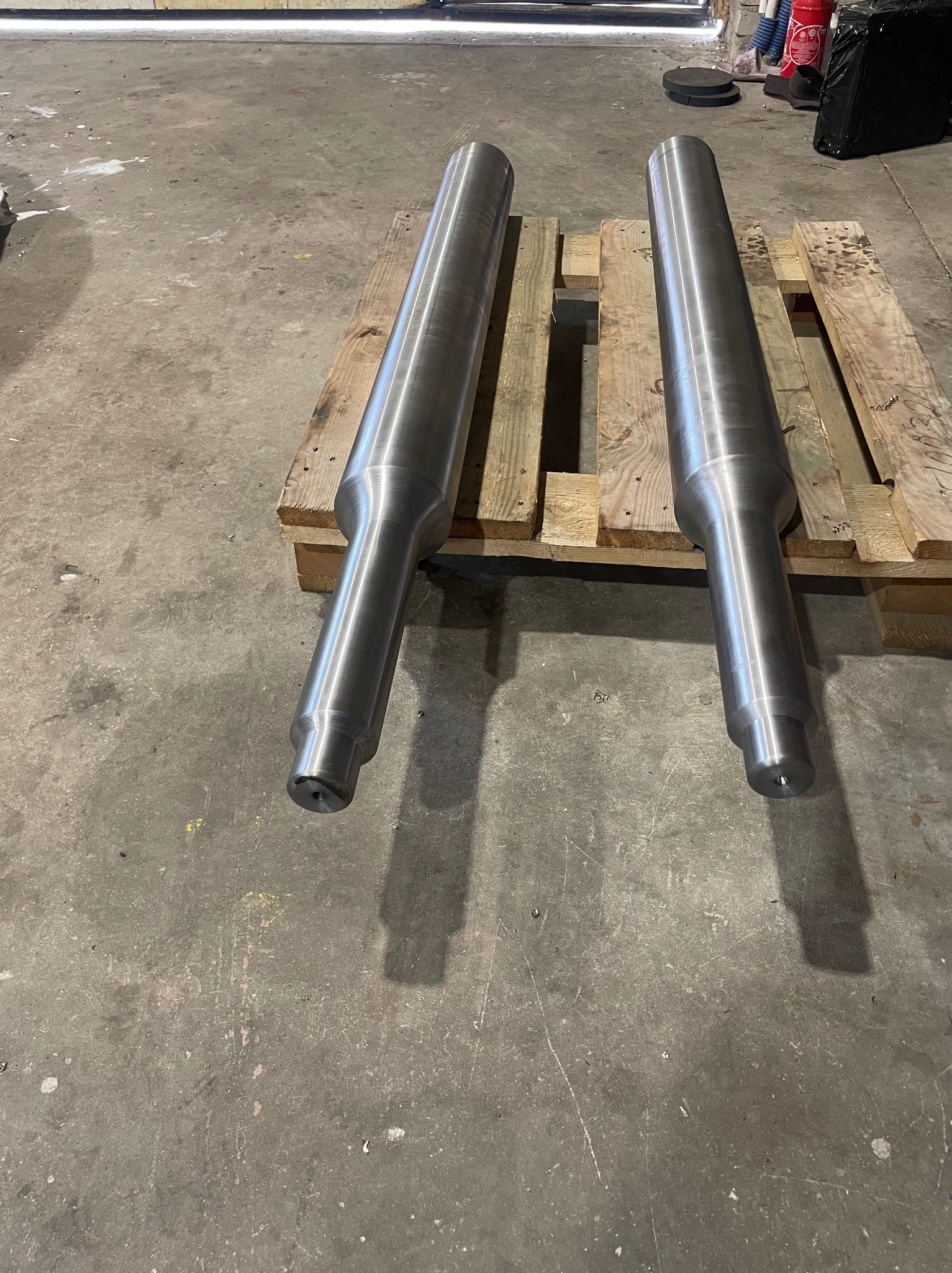 Dwa metalowe, cylindryczne elementy tokarskie na drewnianej palecie w warsztacie. Widoczne ślady obróbki, gładka powierzchnia. Szary beton w tle.