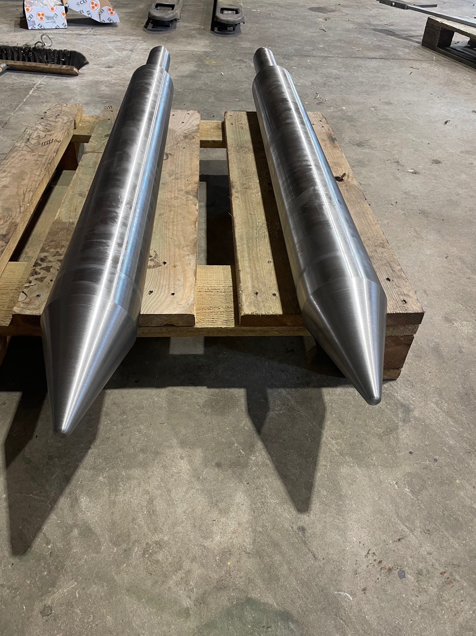 Dwa metalowe, stożkowe elementy na drewnianej palecie, o gładkiej, srebrzystej powierzchni. Widoczne ślady obróbki, surowe tło betonowej podłogi.