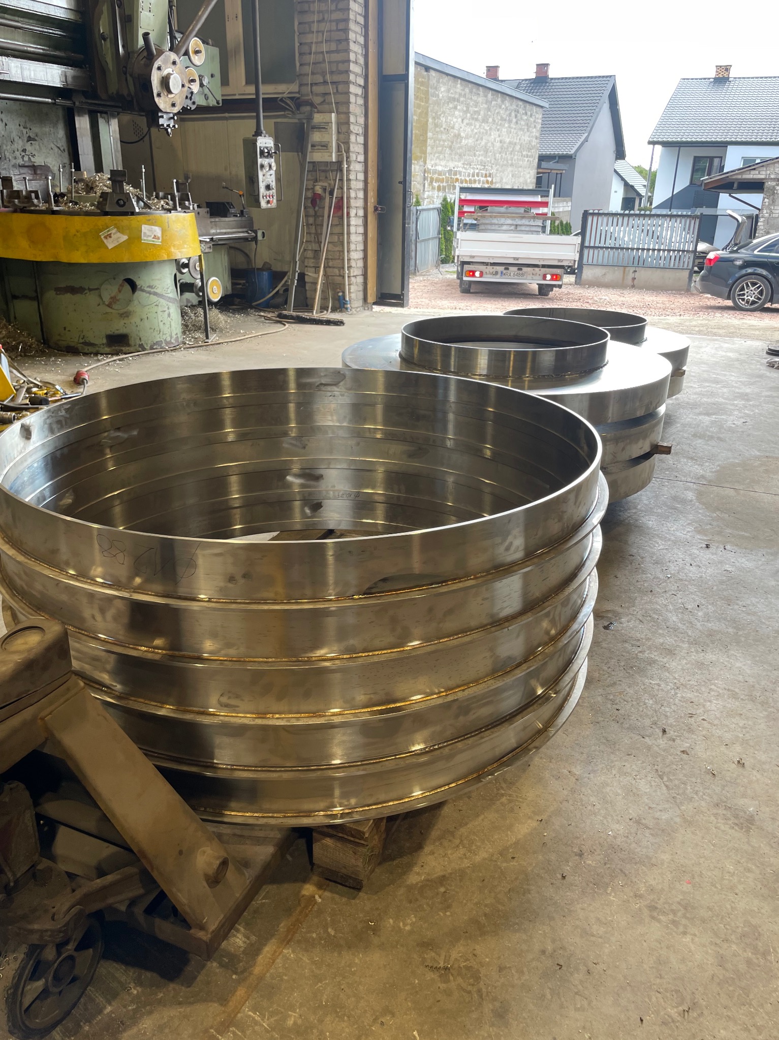 Metalowe pierścienie spawane w Rogowie, ułożone na palecie w warsztacie, w tle tokarka i budynki mieszkalne, co sugeruje produkcję na zamówienie.