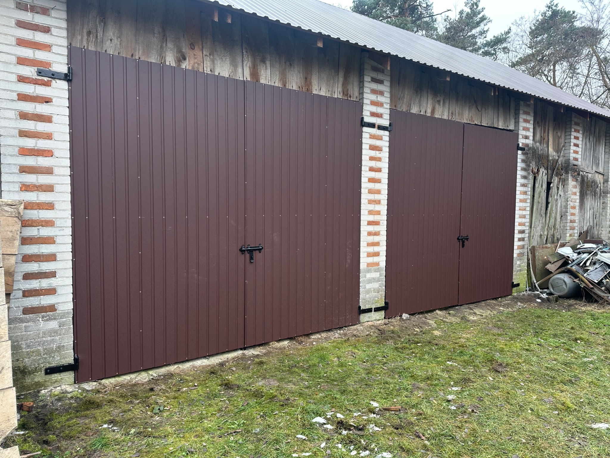 Brązowa, dwuskrzydłowa brama garażowa z blachy trapezowej, zamontowana w murowanym budynku gospodarczym z drewnianą konstrukcją dachu i widocznymi elementami zniszczenia.