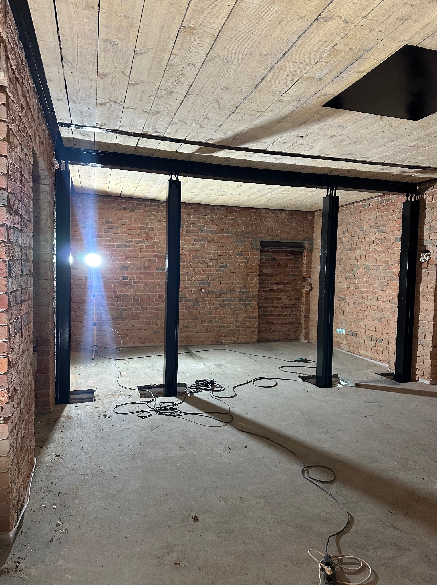 Wnętrze w trakcie montażu stalowej konstrukcji nośnej z czarnych profili na tle ceglanej ściany i drewnianego sufitu. Widoczne kable i lampa budowlana.