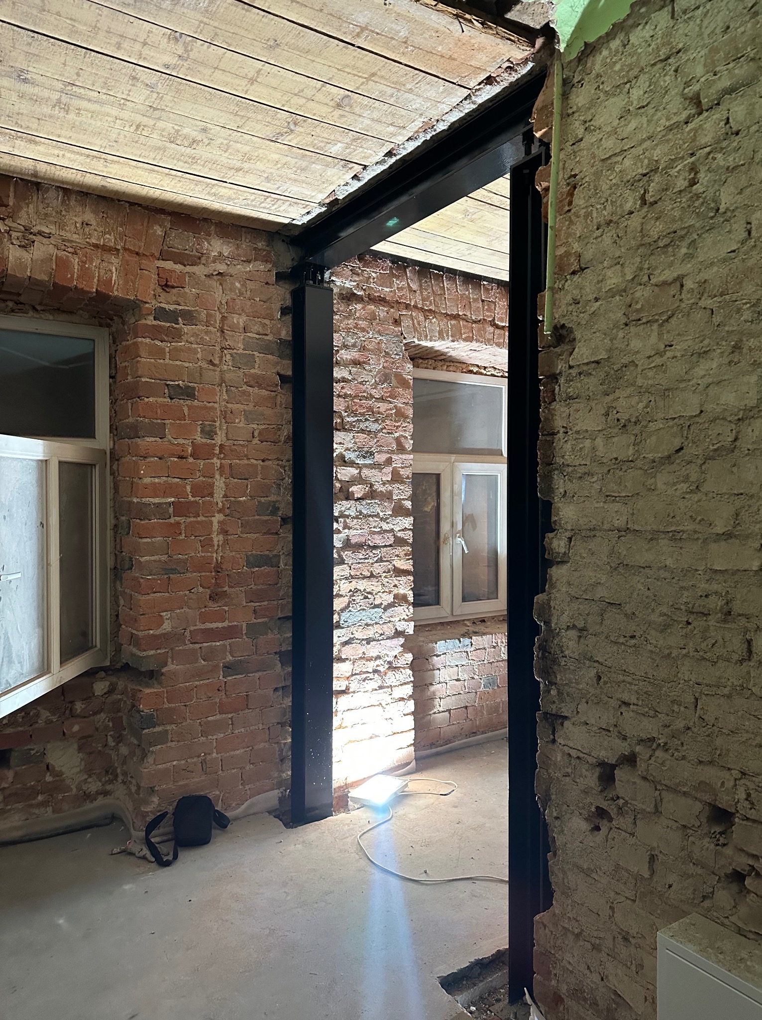 Czarne, metalowe wzmocnienie w surowym wnętrzu z ceglanymi ścianami i drewnianym stropem. Widoczne okno i fragment podłogi. Prace remontowe w toku.