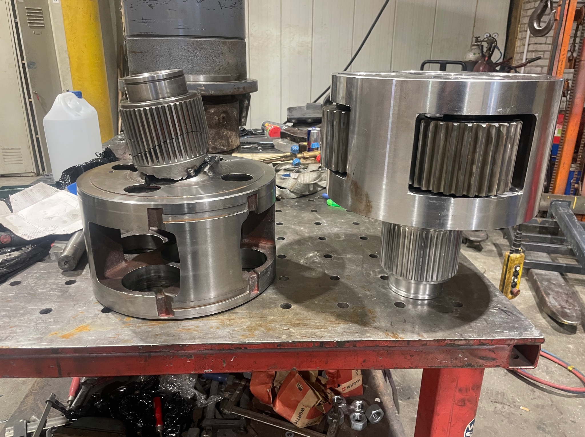 Dwa metalowe elementy z zębatkami na stole warsztatowym. Jeden element uszkodzony, drugi w całości. Widoczne narzędzia i akcesoria warsztatowe w tle.