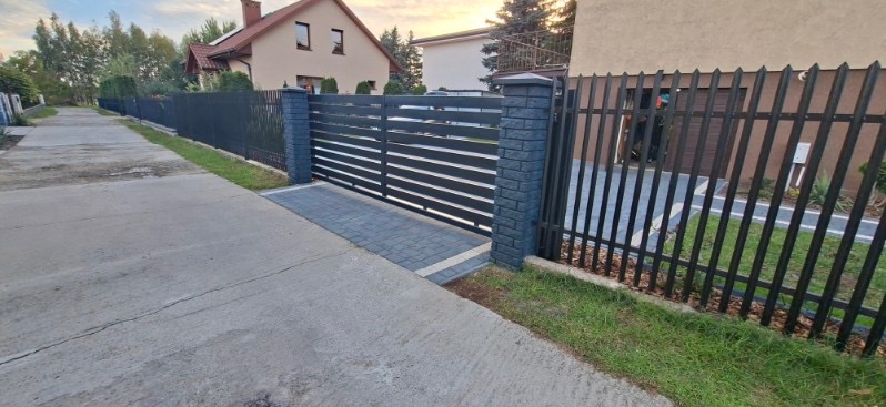 Nowoczesna, pozioma brama wjazdowa z czarnymi przęsłami i murowanymi słupkami, uzupełniona o ogrodzenie panelowe z pionowych prętów. Kostka brukowa na podjeździe.