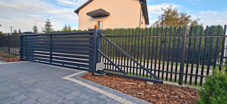 Nowoczesna, przesuwna brama wjazdowa z automatyką, w kolorze grafitowym, z poziomymi panelami, na tle domu jednorodzinnego i ogrodzenia palisadowego.