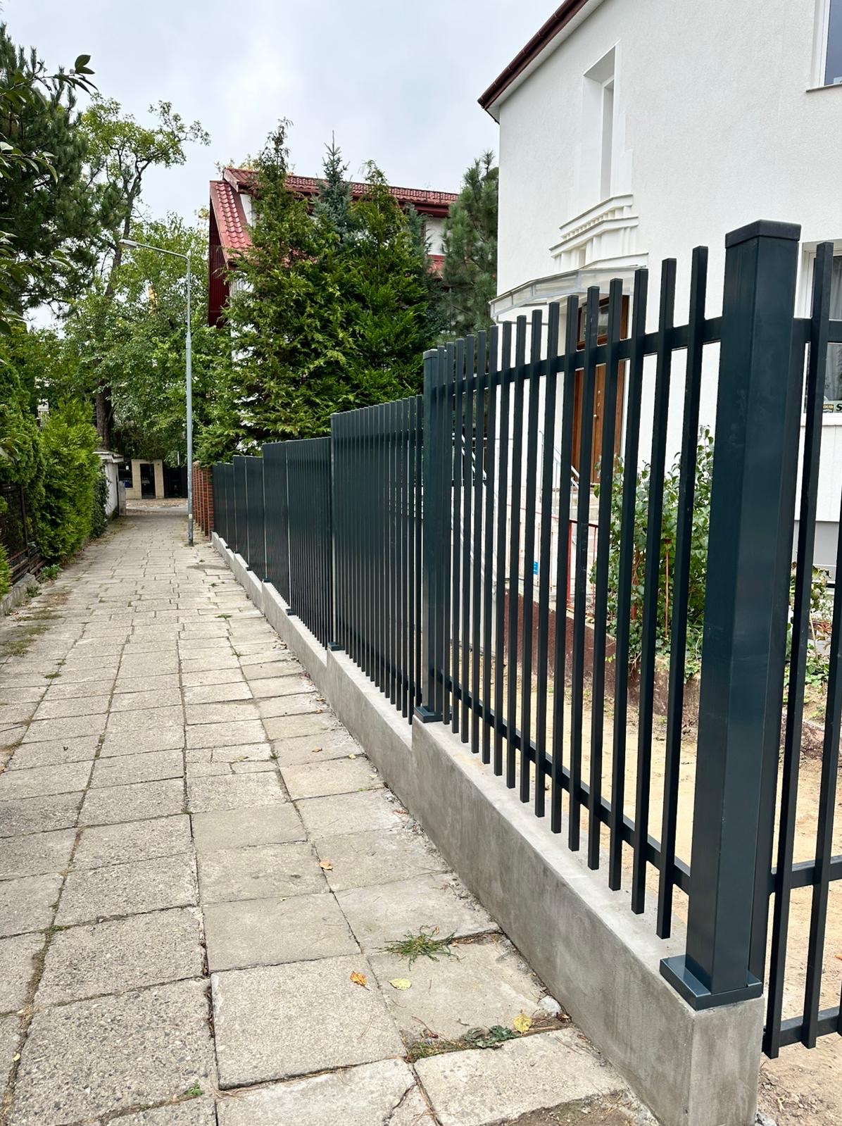Nowoczesne, pionowe ogrodzenie metalowe w kolorze grafitowym, osadzone na betonowej podmurówce, przylegające do chodnika z kostki. W tle dom z czerwoną dachówką.