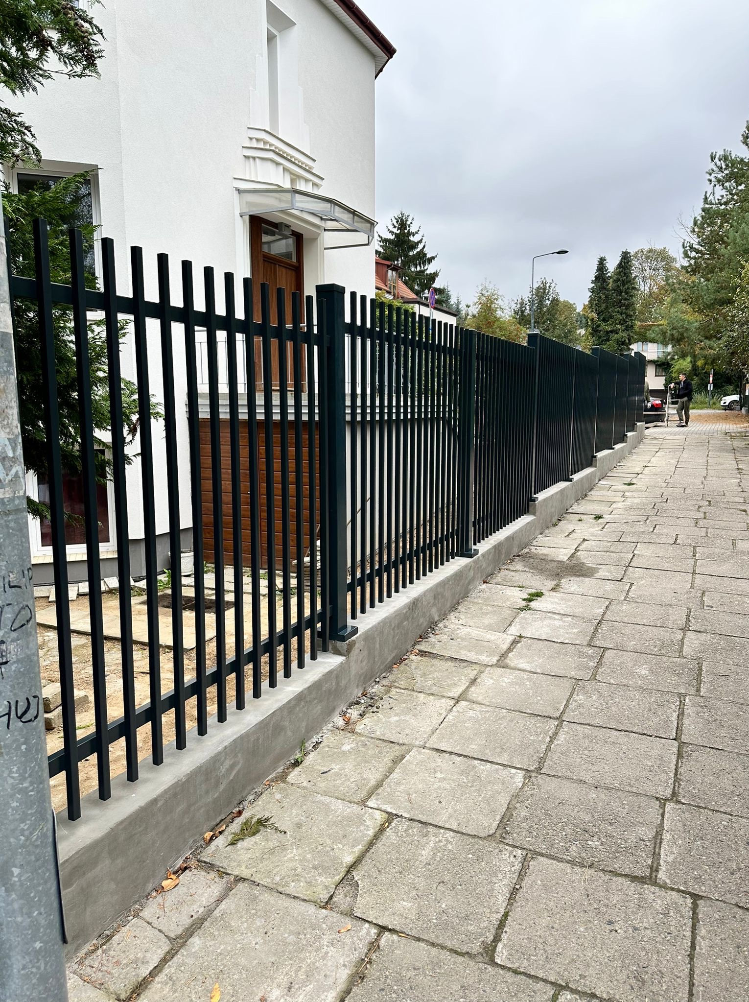 Nowoczesne, czarne ogrodzenie metalowe z pionowymi prętami, osadzone na betonowej podmurówce, przylegające do białego budynku z drewnianymi drzwiami wejściowymi.
