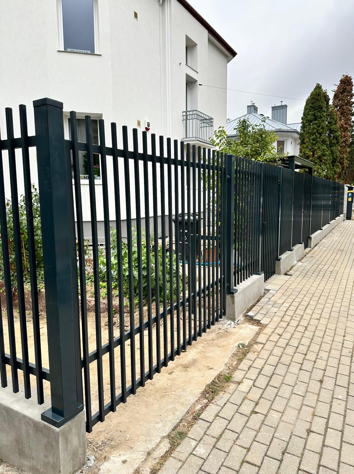 Nowoczesne, metalowe ogrodzenie posesji w kolorze grafitowym, z pionowymi prętami i betonową podmurówką. Widok z boku, na tle białego budynku mieszkalnego.