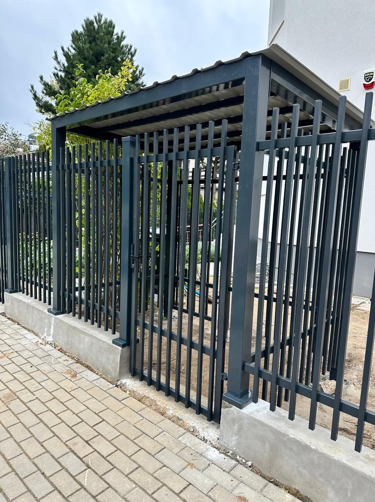 Nowoczesna, grafitowa brama wjazdowa z pionowymi szczeblami i zadaszeniem, osadzona na betonowej podmurówce, widok z zewnątrz na tle kostki brukowej i zieleni.