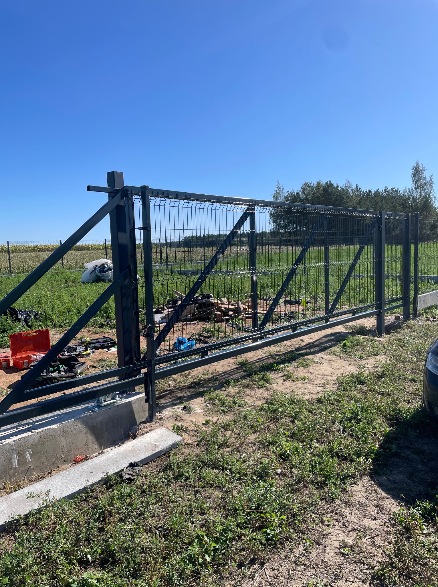 Montaż przesuwnej bramy panelowej na betonowej podstawie. Widoczne narzędzia i elementy konstrukcyjne na tle zieleni i błękitnego nieba. Ujęcie z bliska, w słoneczny dzień.