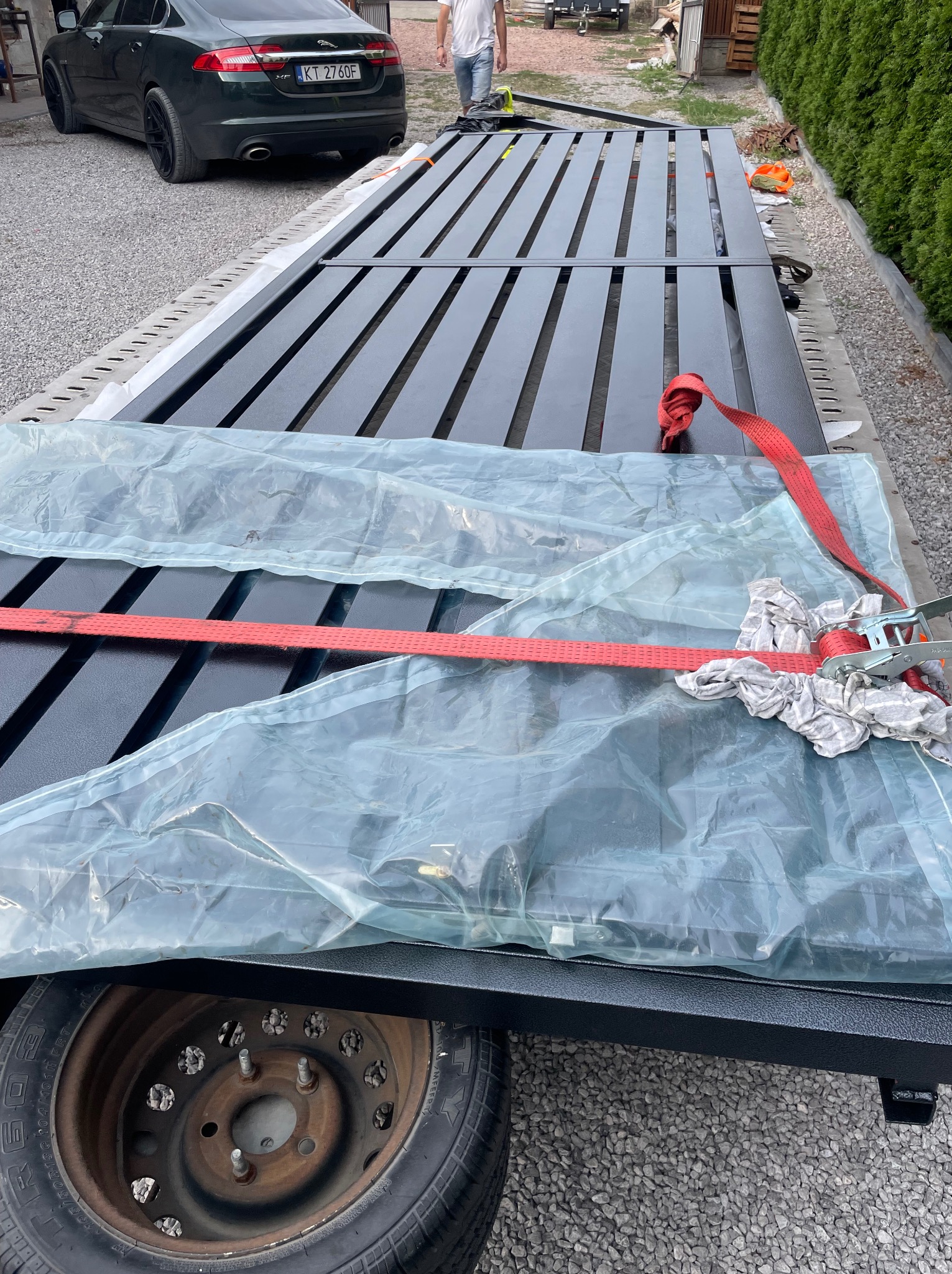 Ciemnoszara, metalowa konstrukcja bramy wjazdowej, zabezpieczona folią i pasami transportowymi, leży na przyczepie. W tle widoczny samochód i fragment zieleni.