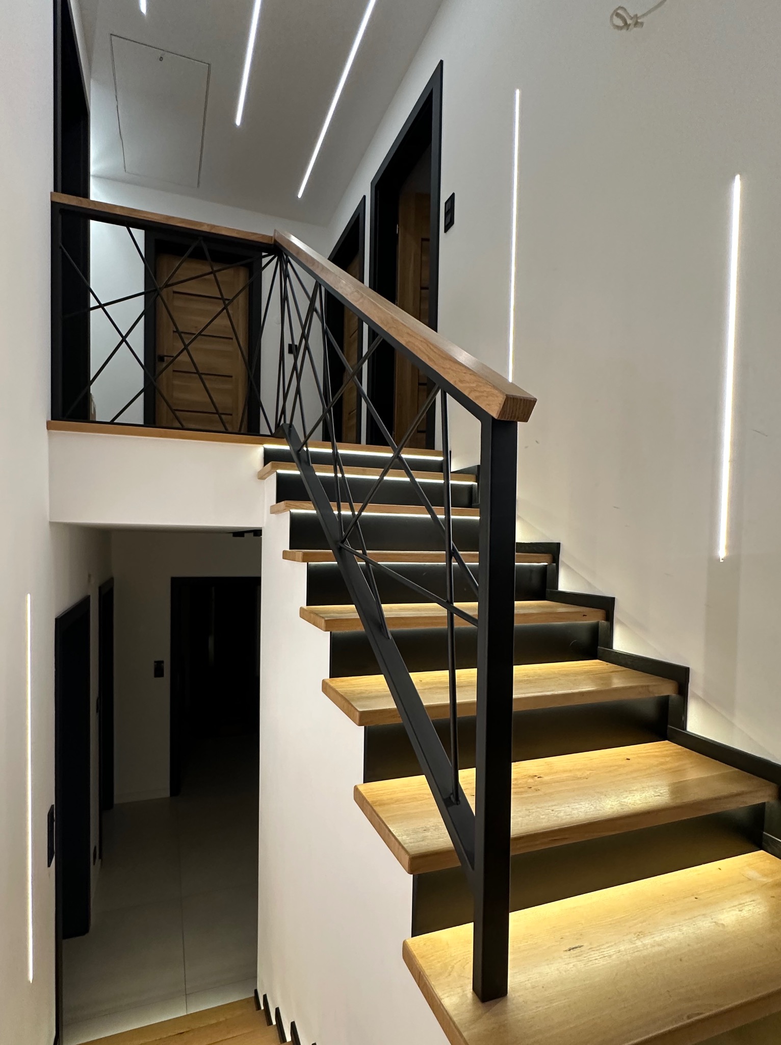 Nowoczesne schody z drewnianymi stopniami i czarną, metalową balustradą o geometrycznym wzorze. Oświetlenie LED podkreśla stopnie i pionowe linie na ścianie.