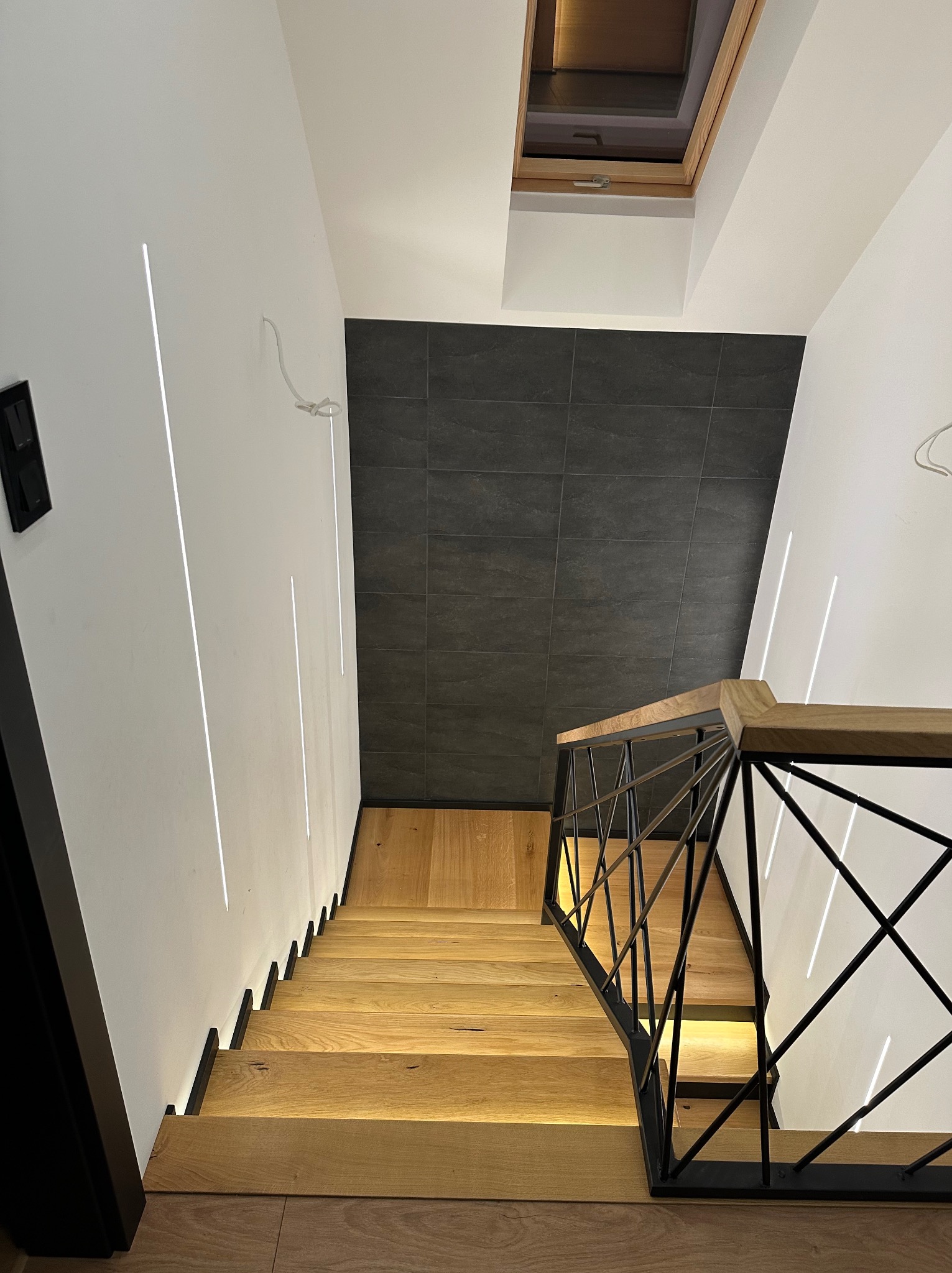Nowoczesne schody drewniane z metalową balustradą w Rogowie. Minimalistyczny design, oświetlenie LED w ścianie, czarne płytki na ścianie. Widok z góry.