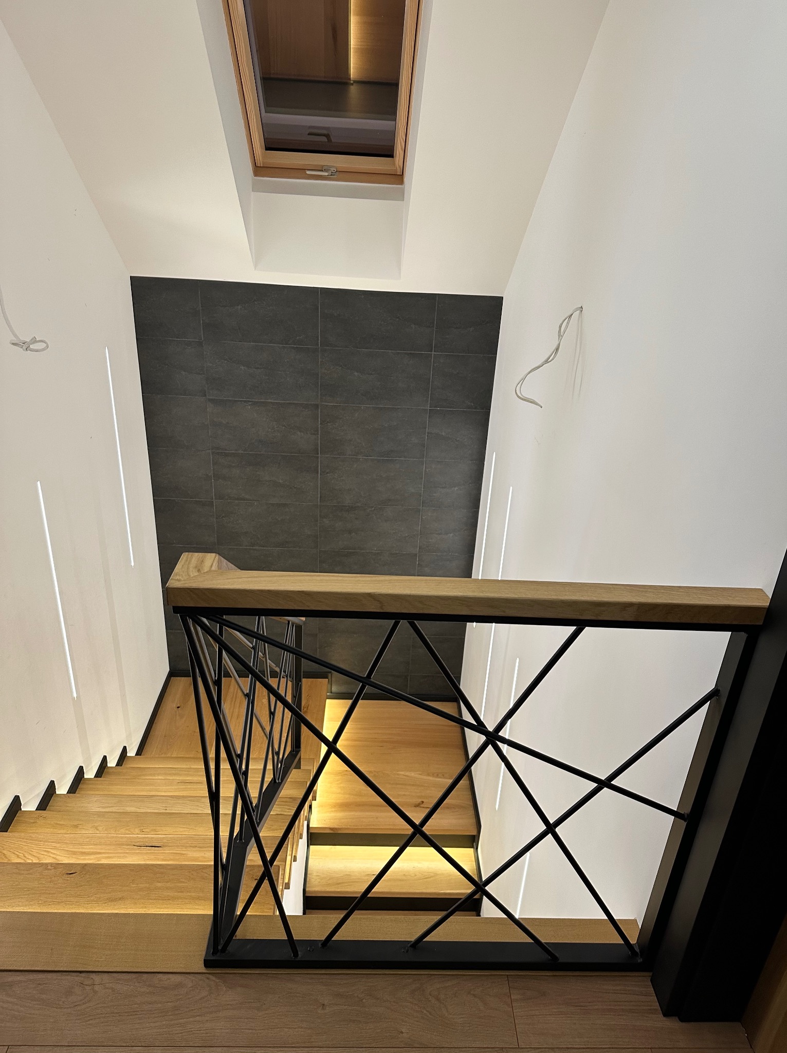 Nowoczesna balustrada schodowa z czarnych prętów i drewnianym blatem, widok z góry na drewniane schody z podświetleniem LED wzdłuż ściany i okno dachowe.