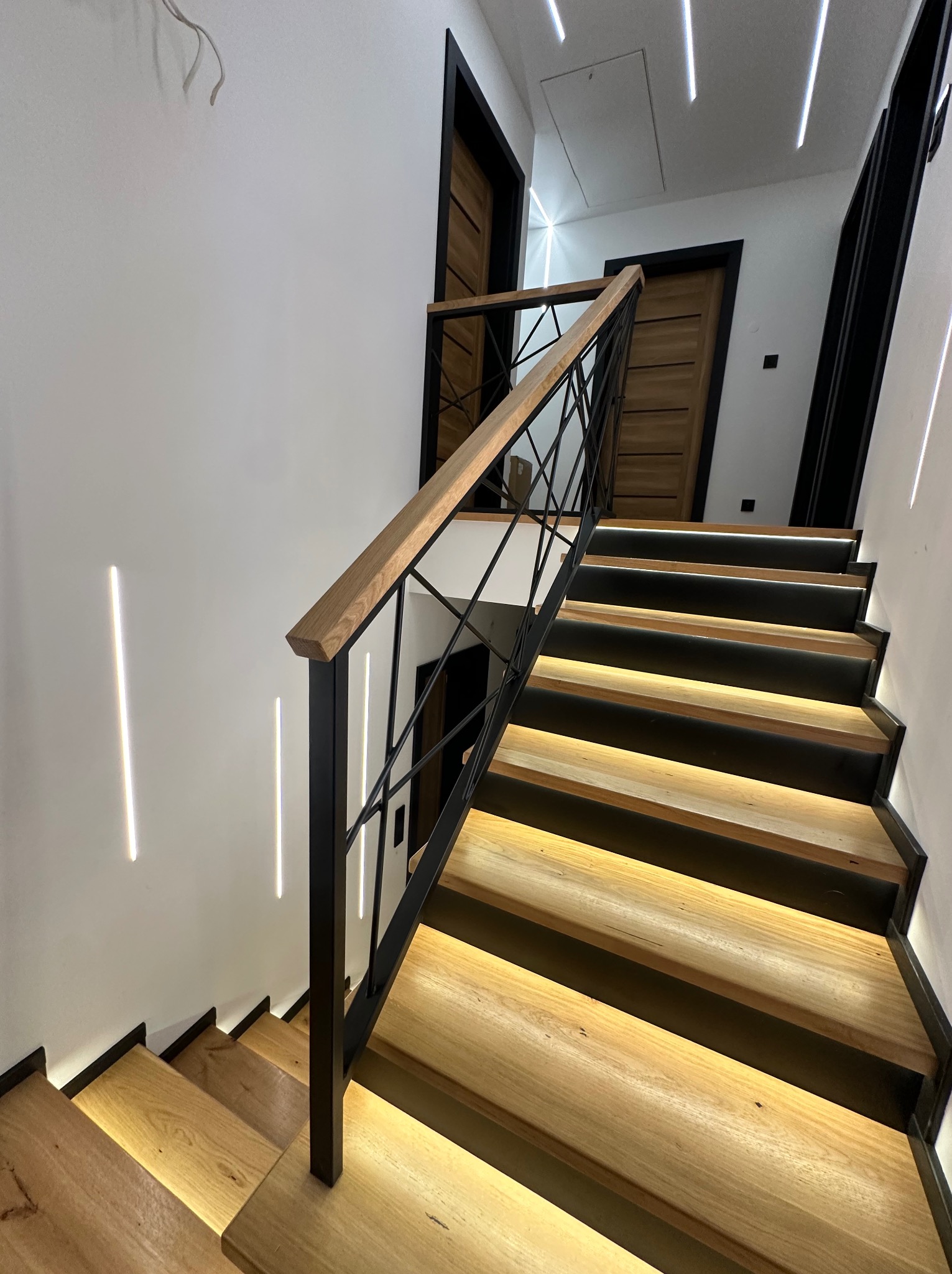 Nowoczesne schody z drewnianymi stopniami i czarną, metalową balustradą o geometrycznym wzorze. Oświetlenie LED podkreśla stopnie i ściany.