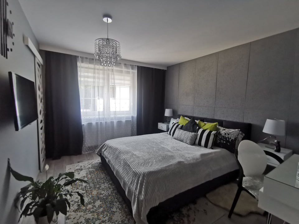 Nowoczesna sypialnia w odcieniach szarości z łóżkiem, dekoracyjnym żyrandolem i ścianą z paneli, akcentowana zielonymi poduszkami.