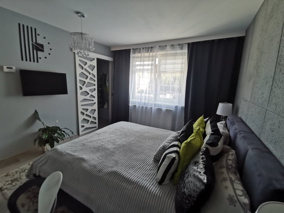 Nowoczesna sypialnia z łóżkiem w szarym odcieniu, dekoracyjną ścianą z paneli, designerskim zegarem ściennym i ażurowymi drzwiami przesuwnymi, oświetlona kryształowym żyrandolem.