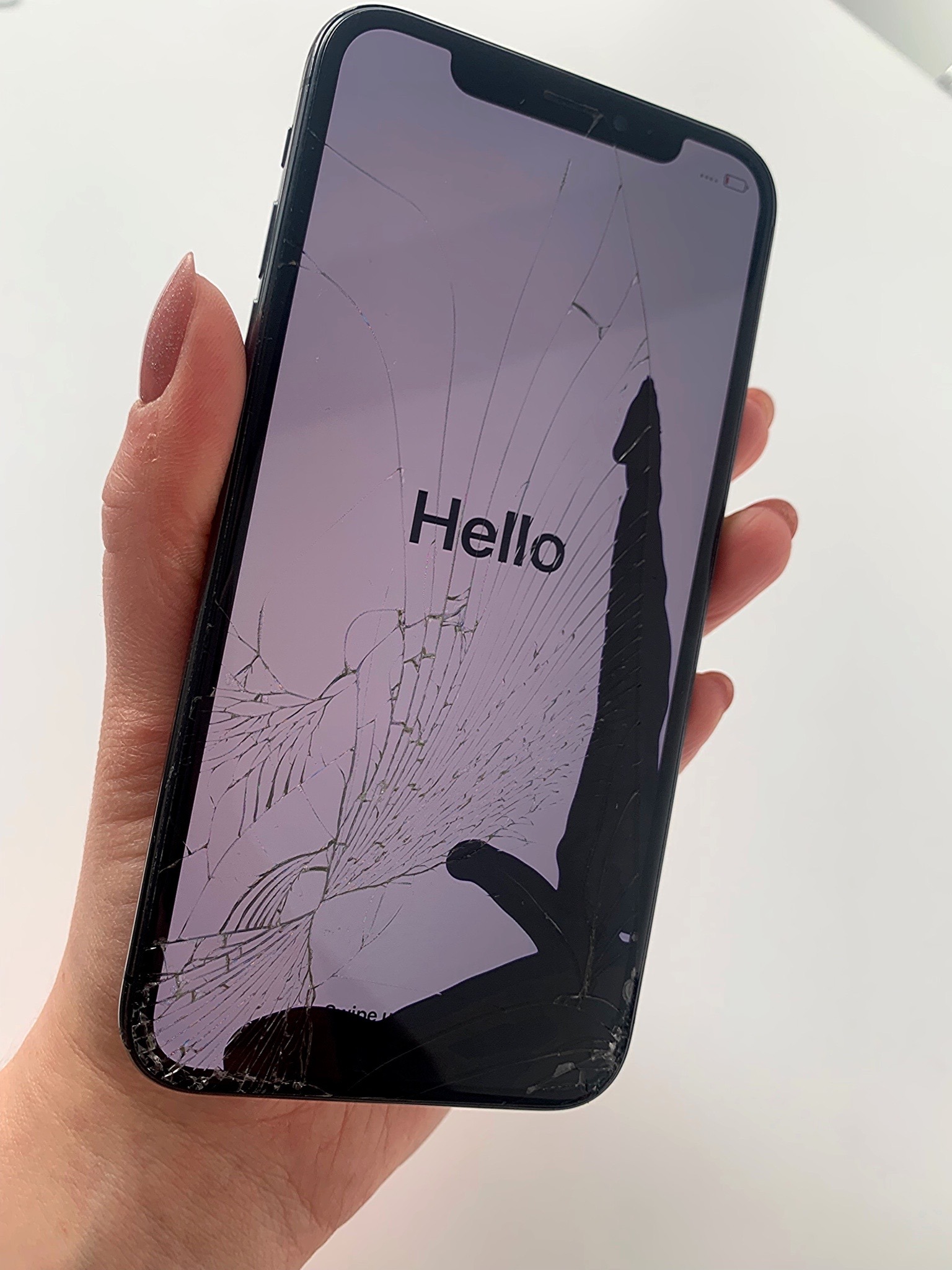 Smartfon z mocno pękniętym ekranem, włączony, trzymany w dłoni na białym tle.