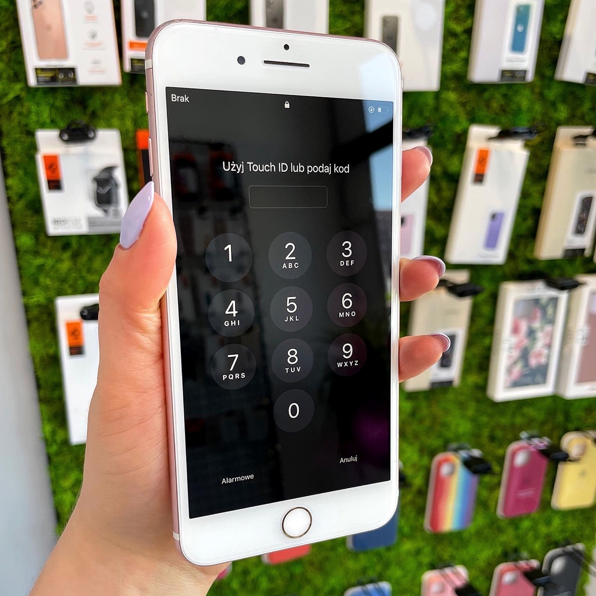 Smartfon zablokowany kodem, trzymany w dłoni z jasnofioletowym manicure, na tle ściany z zielonego mchu i pudełek po akcesoriach do telefonów.