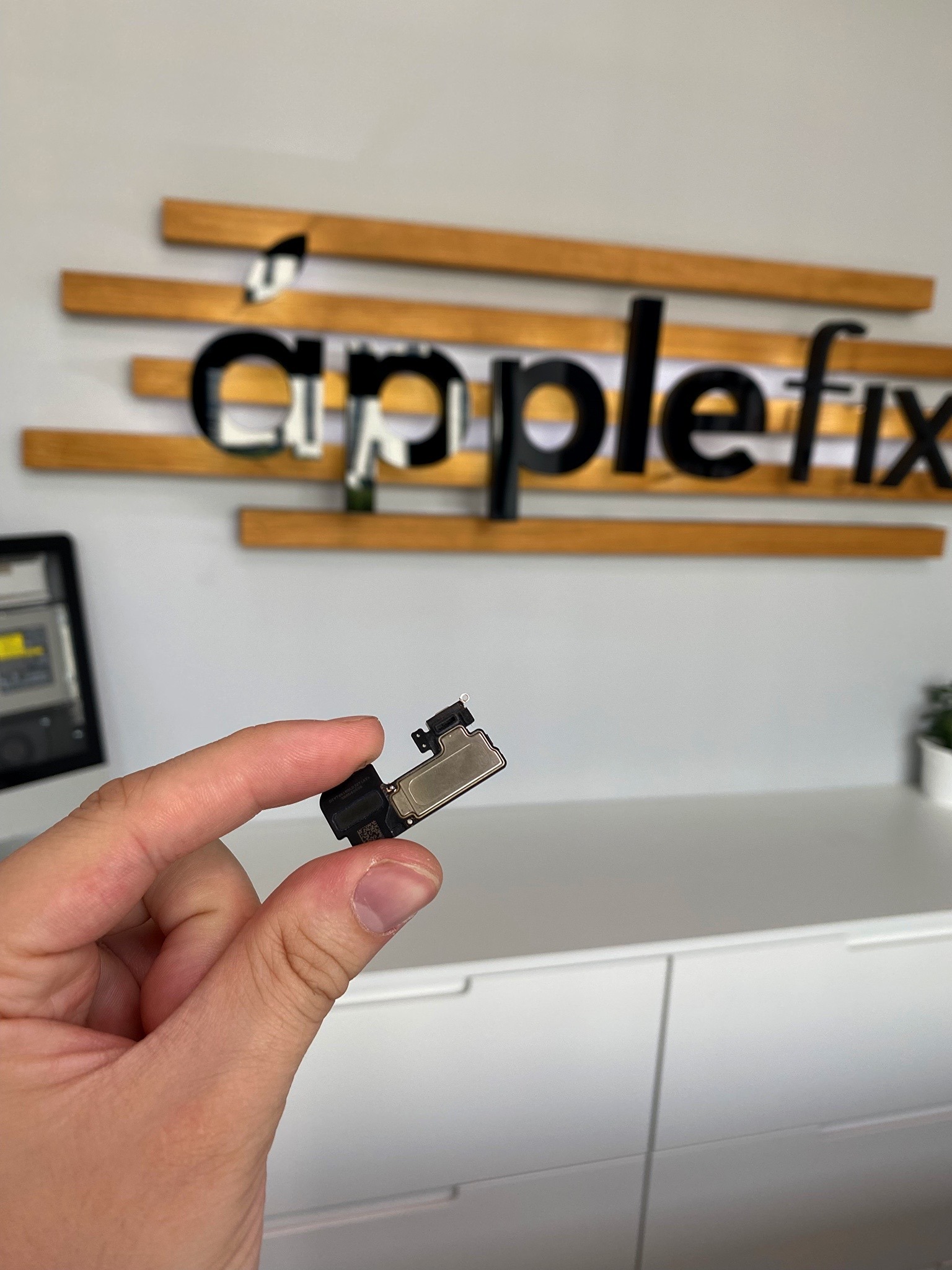 Dłoń trzymająca mały element elektroniczny, prawdopodobnie głośnik lub czujnik iPhone'a, na tle logo 'applefix' i białej komody.