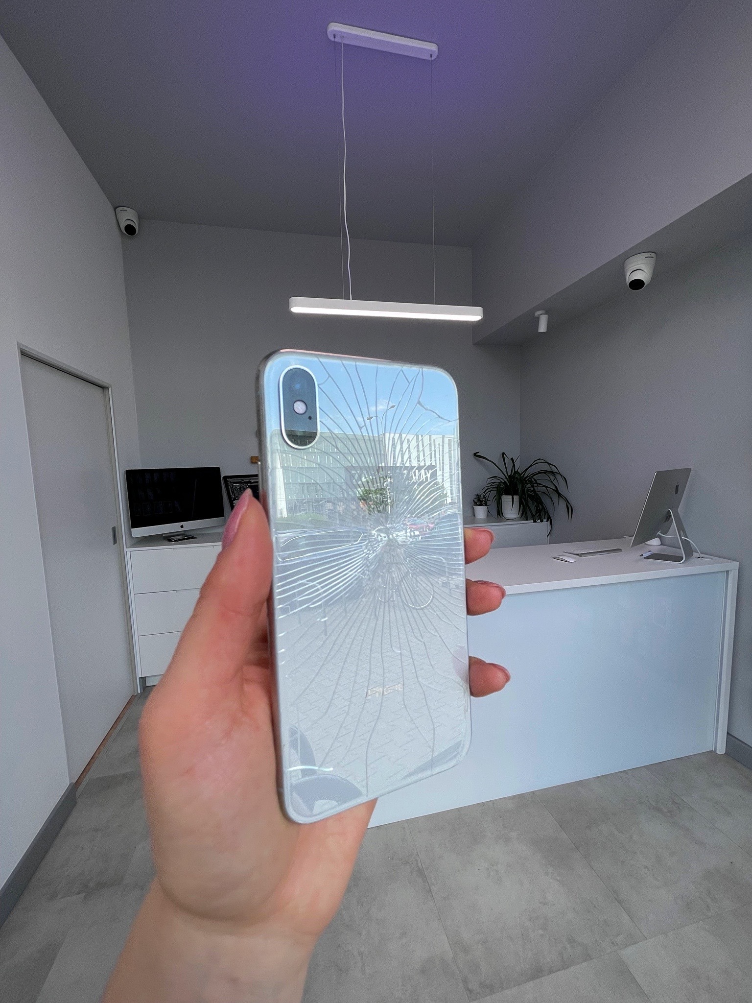 Dłoń trzymająca uszkodzony, srebrny smartfon z pękniętą szybą na tle nowoczesnego biura z białymi meblami i monitoringiem.